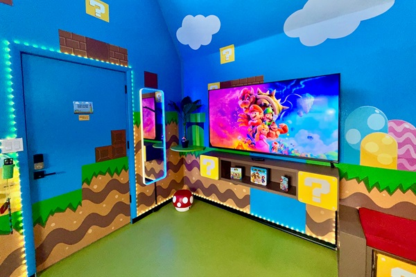 Vibrant blue walls with Super Mario murals, glowing question mark blocks, green pipes, and brick details surround the extra large 75" smart TV (+ Nintendo Switch system). The mirror lights up and plays music via Bluetooth, perfect for family fun!