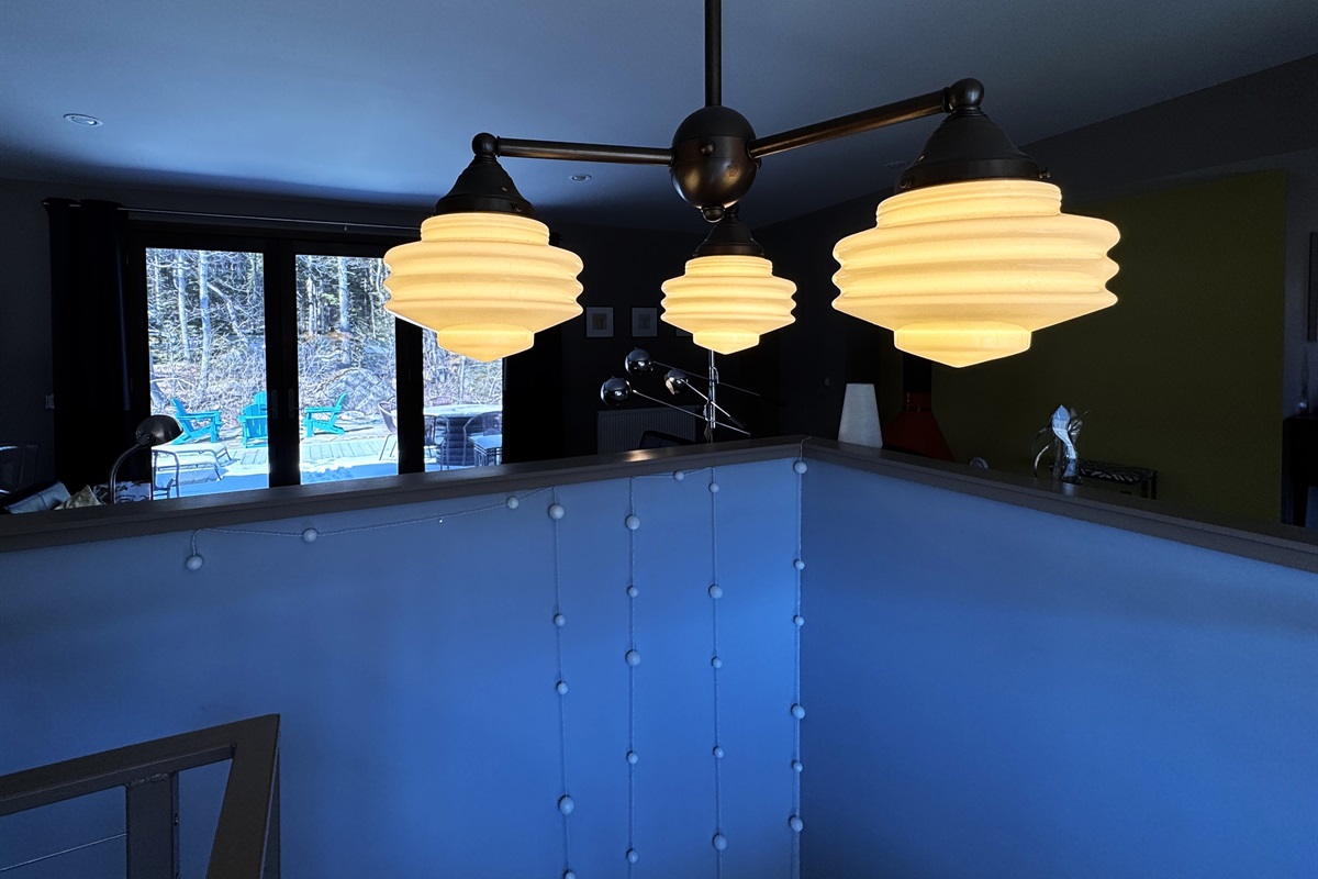 Modern designer lighting over the main stairway, framed by large picture windows and a direct-access door to the 33ft "Park Border" deck. This bright transition offers a seamless indoor-outdoor flow at the edge of Acadia National Park.