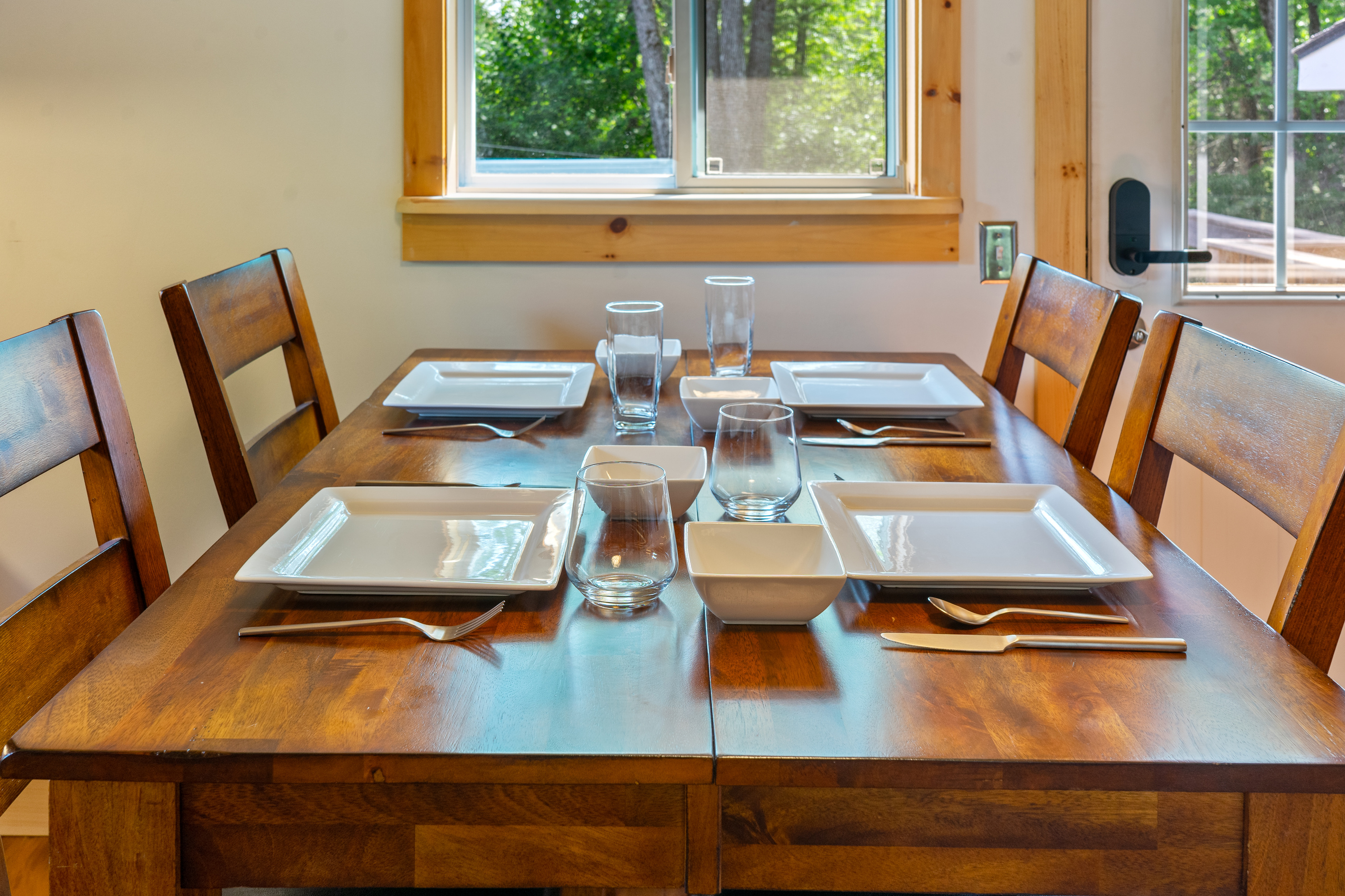 Enjoy family meals at the wooden dining table, beautifully set with contemporary square dinnerware, glassware, and flatware. This comfortable dining space sits adjacent to a bright slider window and the main entry door leading out to the deck.