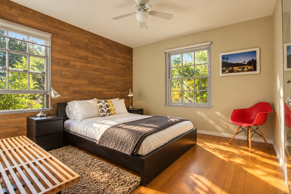 A bright bedroom with a wood feature wall, white bedding and a textured throw. Two windows bring in greenery and sunshine, while a red accent chair, slatted wood bench, bedside table and ceiling fan complete the room.