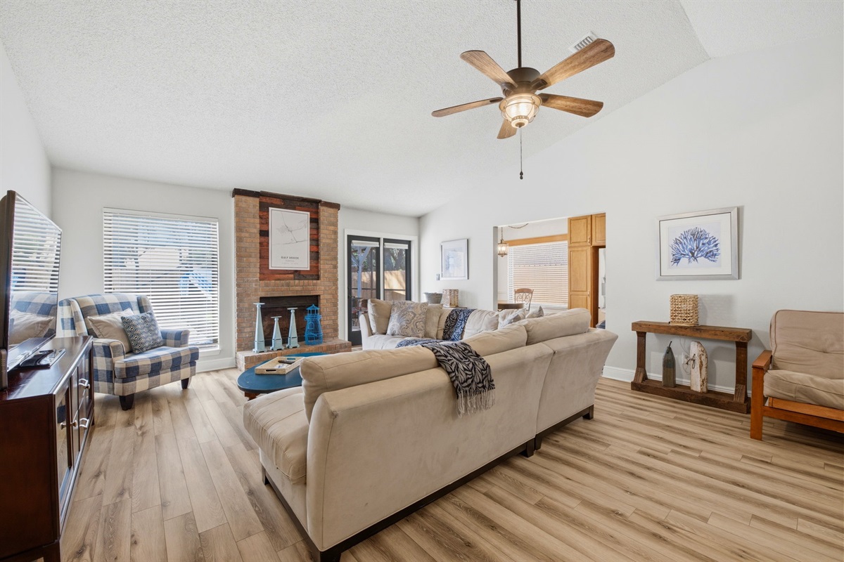 Relax in this bright and inviting living room featuring vaulted ceilings, warm wood flooring, and comfortable seating. A brick fireplace adds charm to the space.  The open layout makes it perfect for gathering, watching TV, or unwinding after a long day.