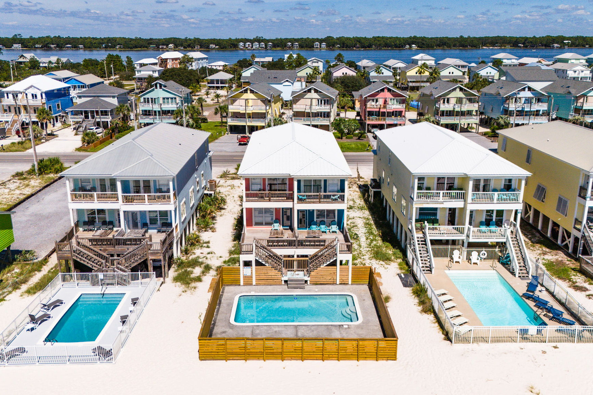 Drone aerial view showcasing multiple colorful beach homes, pools, spacious outdoor decks, and the surrounding coastal neighborhood.