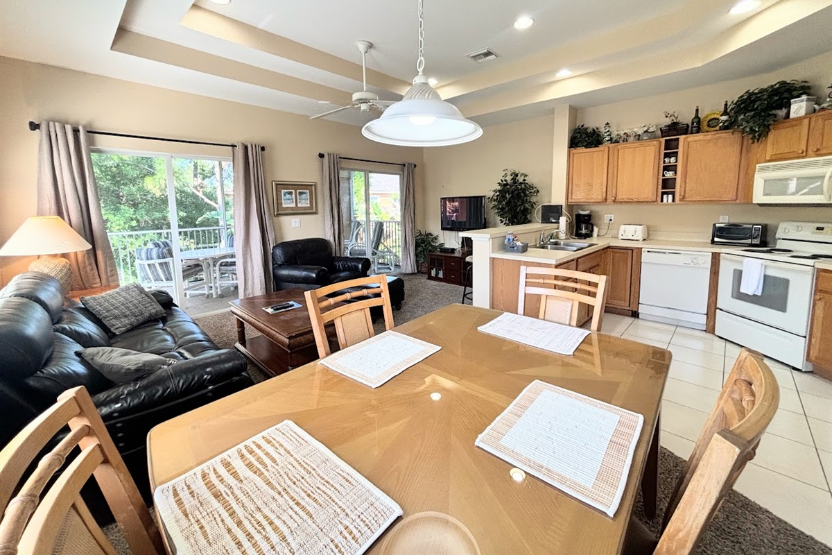 Bright and spacious open-concept living area featuring vaulted tray ceilings, a fully equipped kitchen, comfortable seating, and sliding glass doors leading out to a private screened balcony.