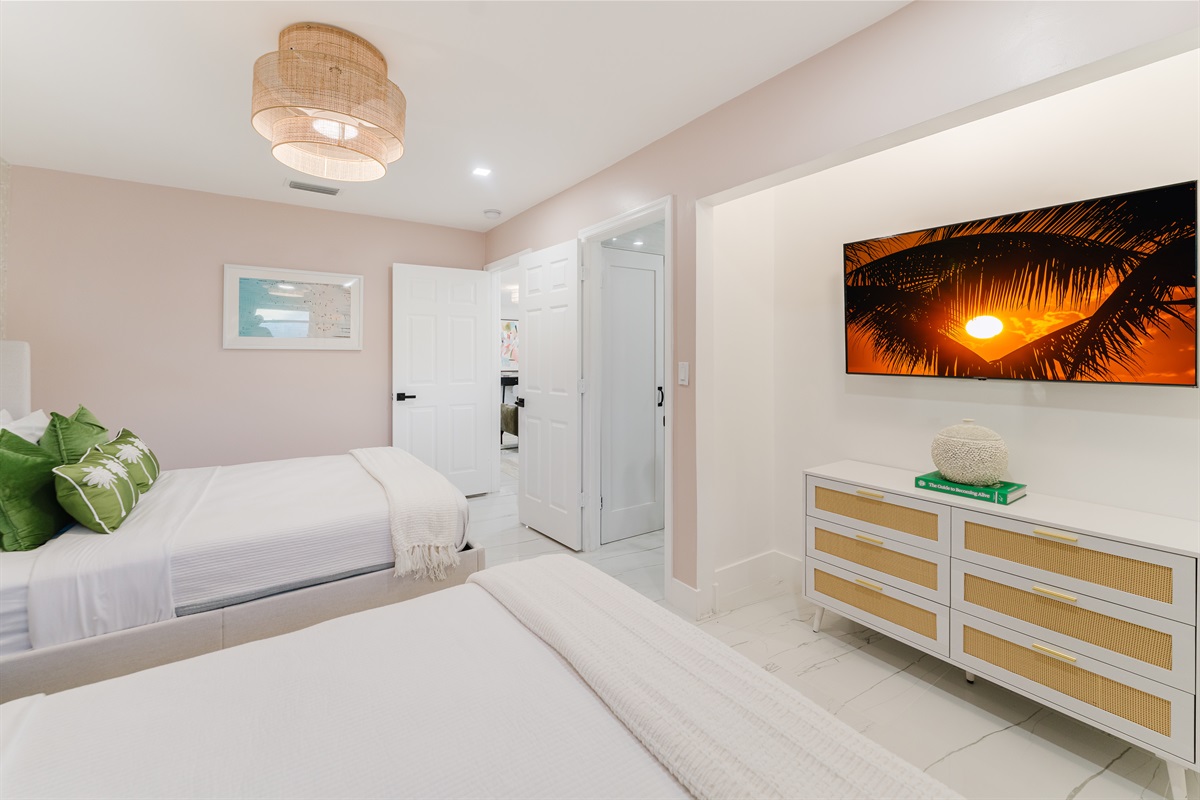 Bright, serene guest suite with two queen beds, hotel-quality linens, a wall-mounted smart TV, and ample storage. Features blackout curtains and blinds, a rattan ceiling fan, and a private en-suite bathroom—ideal for families or guests sharing space.