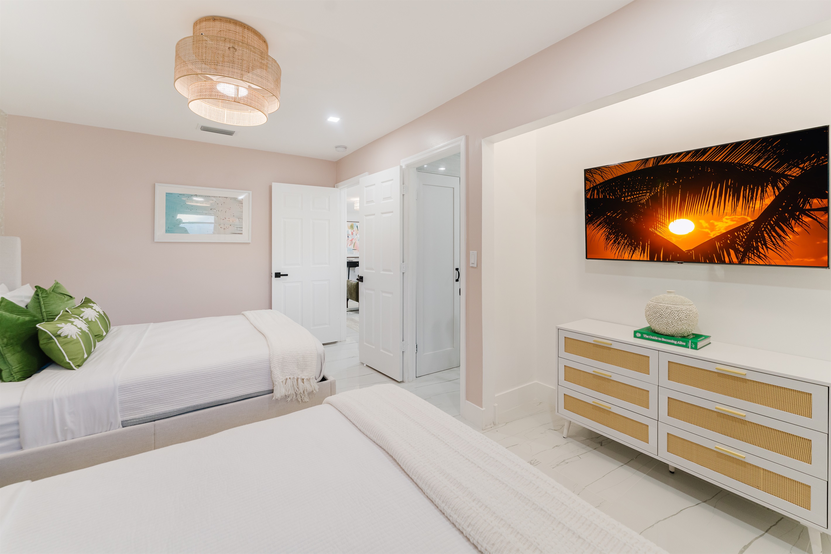Bright, serene guest suite with two queen beds, hotel-quality linens, a wall-mounted smart TV, and ample storage. Features blackout curtains and blinds, a rattan ceiling fan, and a private en-suite bathroom—ideal for families or guests sharing space.