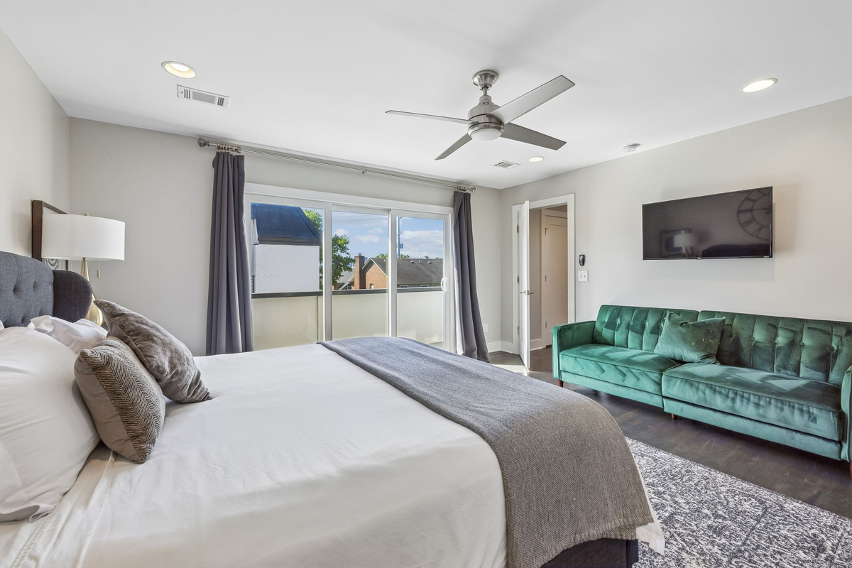 Emerald green tufted sofa adds a vibrant pop of color to this spacious bedroom lounge - Wall mounted TV and large windows make this a great spot for relaxing or streaming - Sophisticated black out drapes and detailed rug complete the room&rsquo;s upscale feel