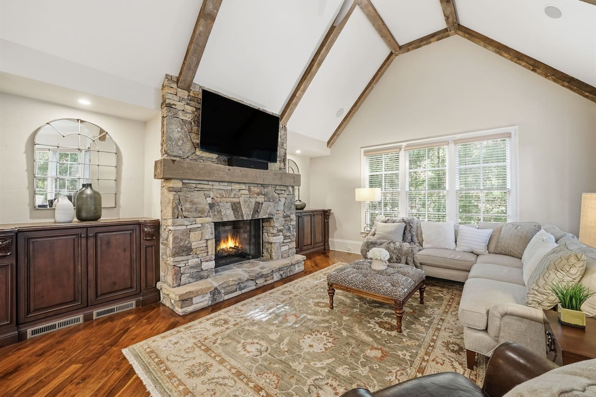 Relax in a warm and welcoming living room featuring high ceilings, a beautiful stone fireplace, and rich wood accents that create a cozy, home-like atmosphere. Enjoy a smart TV with a quality sound system perfect for movie nights and quiet mornings.