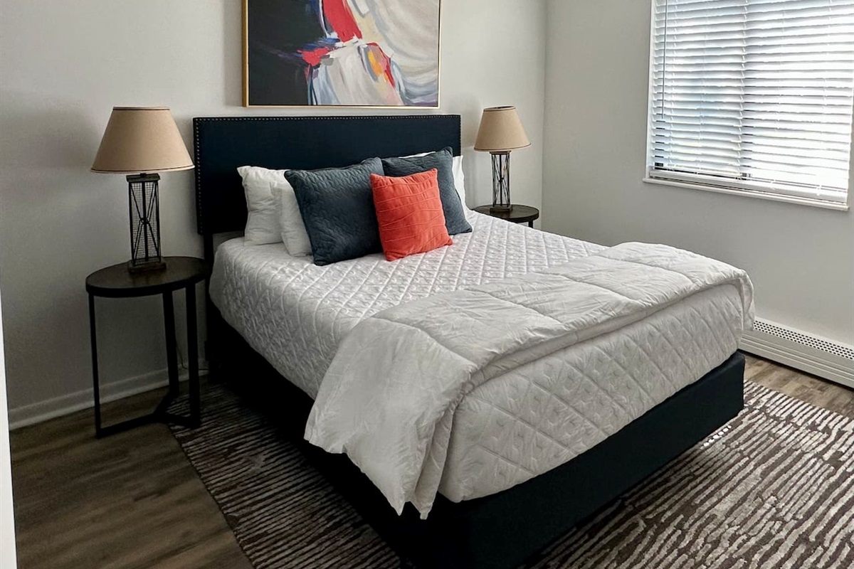 Rest easy in this bright, modern bedroom featuring a plush queen bed with cozy linens and colorful accents. A large mirror, natural light, and calming decor create a peaceful retreat—perfect for a great night’s sleep during your short-term stay.