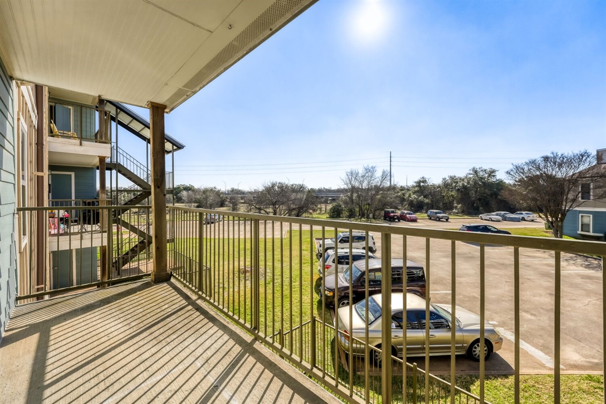 Sunny balcony with a view of the parking area and open green space.