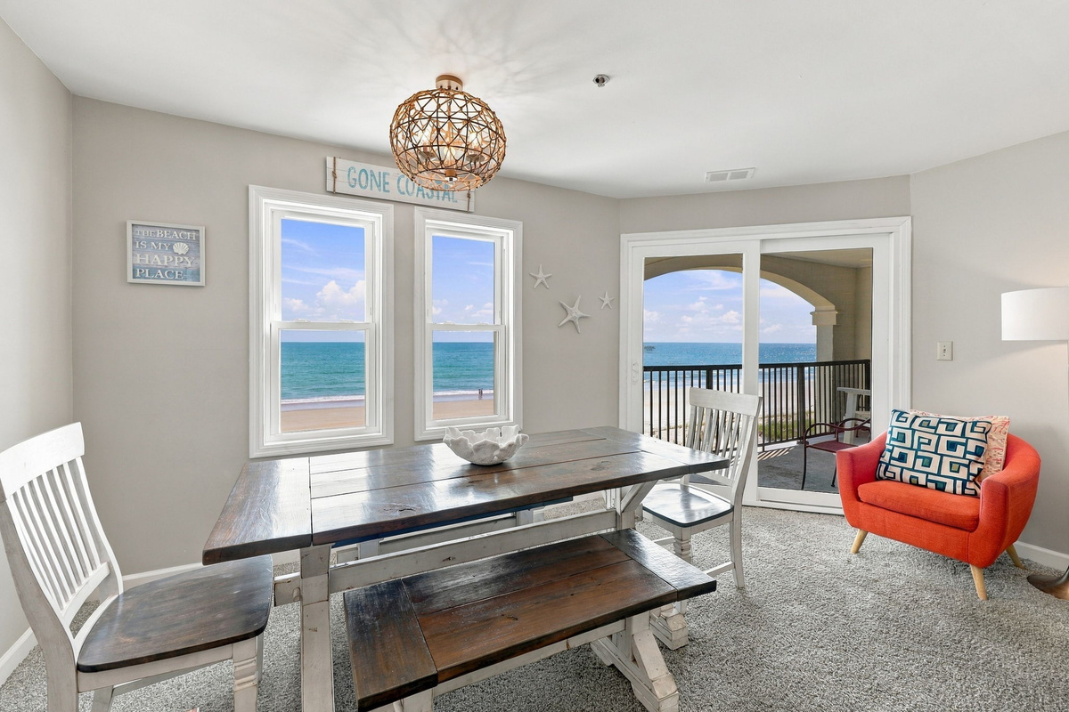 Coastal dining nook anchored by a rustic wooden table with bench seating, woven orb chandelier, and expansive ocean views framed by large windows and glass balcony doors.   
