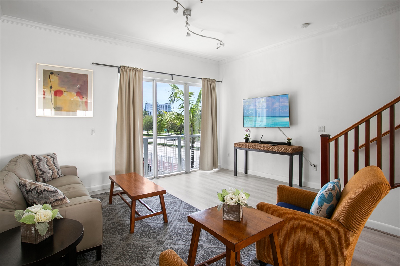 Living room with leather sofa, accent chairs, and a 50-inch flat screen TV. Sliding glass doors open to a Juliet balcony for sea breeze.