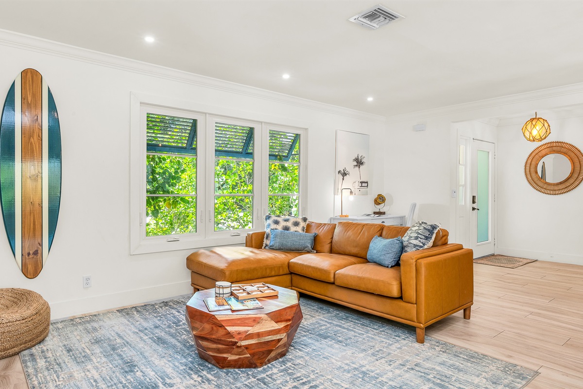 The bright and airy living room features a modern leather sectional, warm wood accents, and lush greenery views through large picture windows. Stylish touches like the surfboard décor and geometric coffee table create a relaxed coastal vibe.