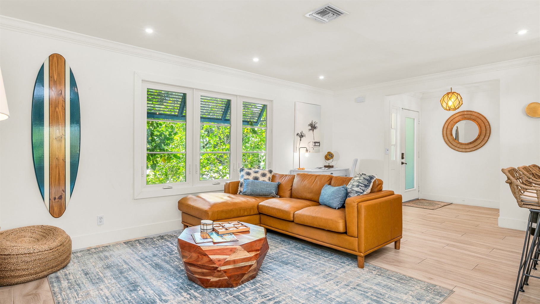 The bright and airy living room features a modern leather sectional, warm wood accents, and lush greenery views through large picture windows. Stylish touches like the surfboard décor and geometric coffee table create a relaxed coastal vibe.
