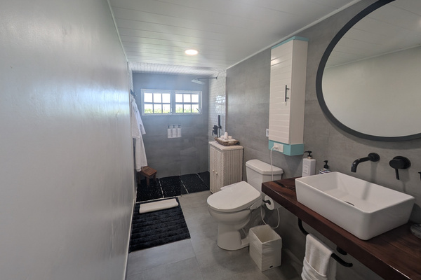 Modern upstairs bathroom at Villa Coral featuring a wood vanity with a white vessel sink, matte black wall-mounted faucets, large round mirror, concrete-gray tiles, and a walk-in shower in Dorado, Puerto Rico.
