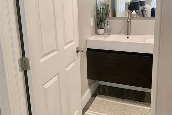 This sleek half-bath features a contemporary floating vanity with a clean white countertop and a minimalist faucet. The modern design is accented by a simple rug and a touch of greenery, providing a stylish and convenient space for guests.