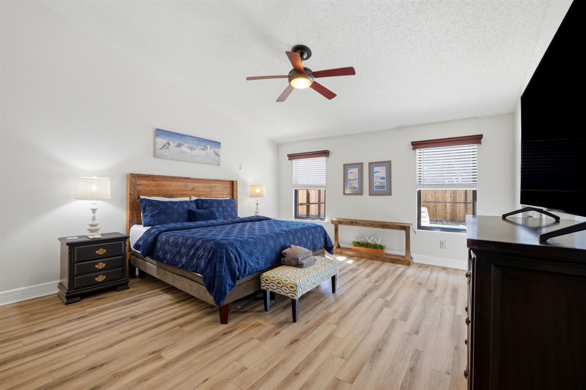 Designed for comfort and space, this bedroom features a plush king bed, stylish furnishings, and warm wood flooring throughout. Large windows bring in natural light, while a ceiling fan keeps the room cool and inviting.