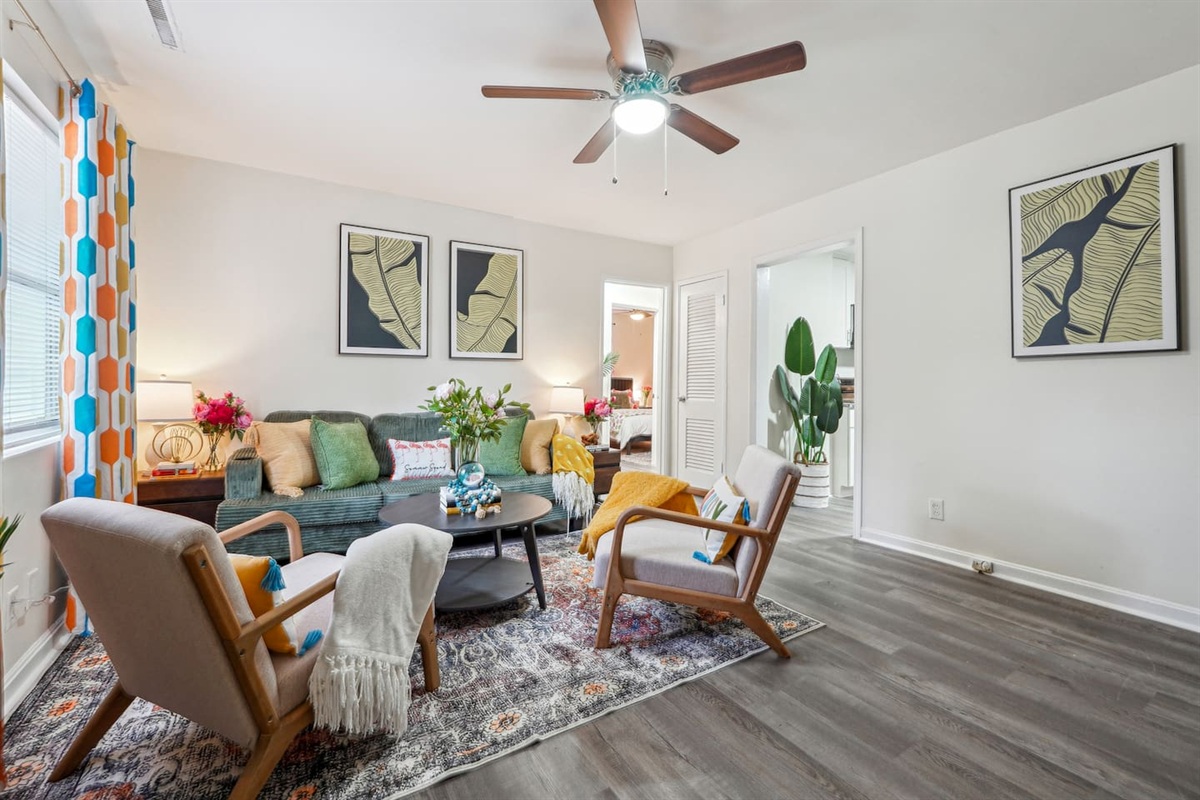 Enjoy a cozy living room featuring a long comfortable sofa, a TV for your entertainment, a ceiling fan, two chair, and a window with a lovely view outside — perfect for relaxing and unwinding.
