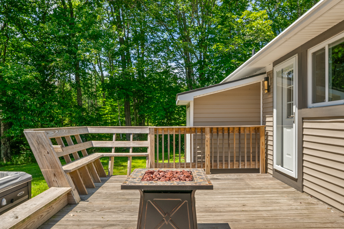Gather on the spacious outdoor wooden deck featuring a central propane fire table filled with lava rock. Relax on the wrap-around integrated wooden bench seating located right outside the glass-paneled rear entry door to the kitchen area.