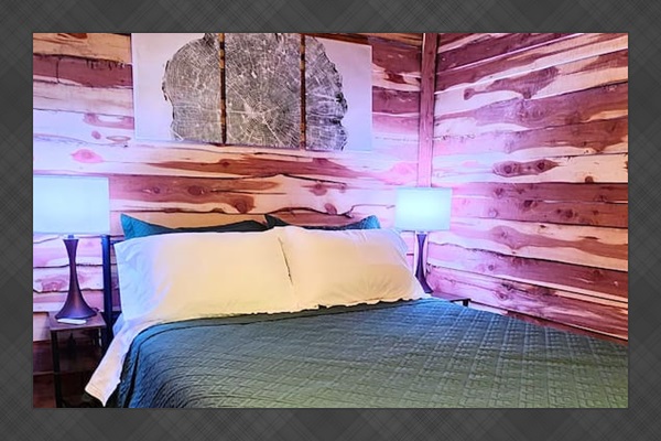 This cozy bedroom features warm cedar walls, a braided rug, and a plush queen bed with a deep green quilt. Soft lighting and a striking tree-ring wall art create a calming, nature-inspired space perfect for unwinding after a day outdoors.