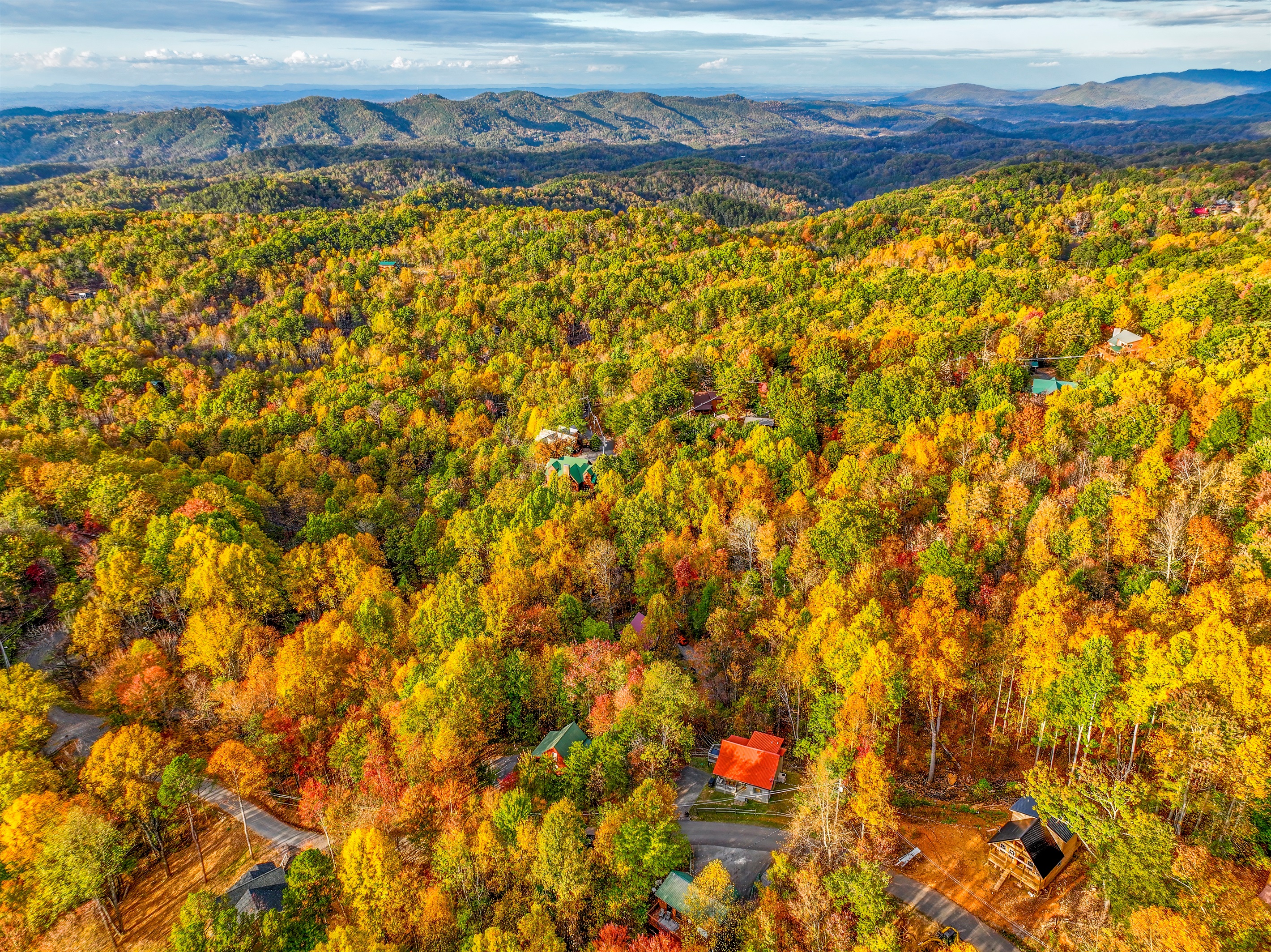An aerial view of a serene autumn landscape, where vibrant hues of gold, orange, and red blanket the forest. Cozy cabins nestle among the trees, with majestic mountains rising in the distance.