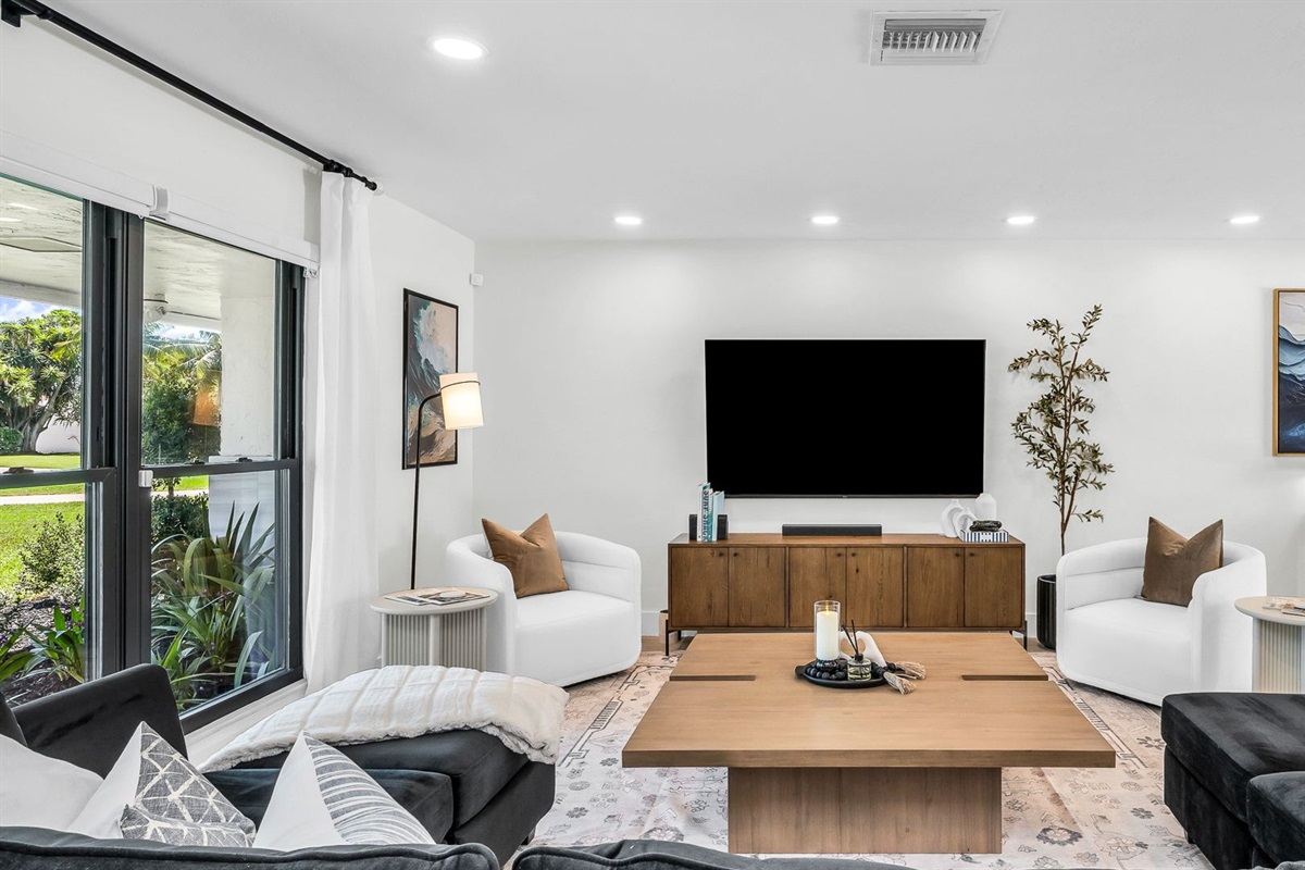Elegant open-concept living room featuring a plush black velvet sectional, 75” smart TV, and designer mid-century modern furnishings. Floor-to-ceiling windows and French doors fill the space with natural light and connect seamlessly to the outdoor pool.