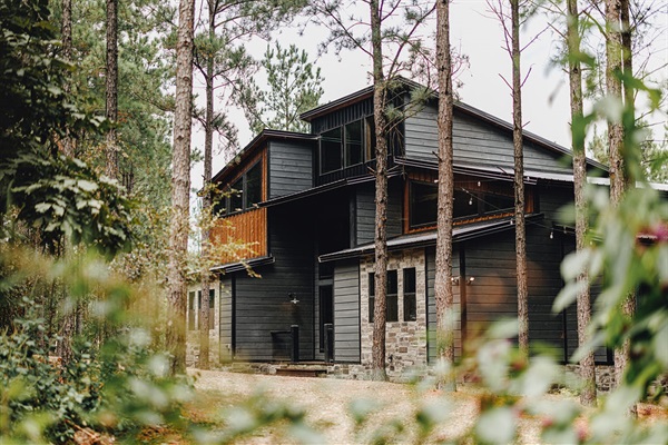 A stunning modern cabin nestled among tall pine trees, featuring a dark wood exterior with large windows. This luxurious retreat is designed for both relaxation and adventure, offering a tranquil escape into nature.