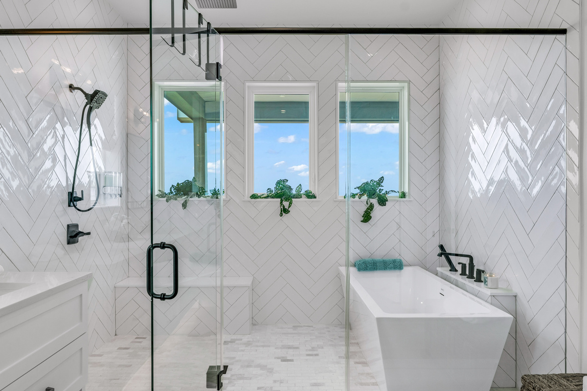 Cair Paravel: The primary bath delivers true spa energy: a freestanding soaking tub beneath windows framing marsh views, a glass rain shower, and dual vanities wrapped in floor-to-ceiling herringbone tile.