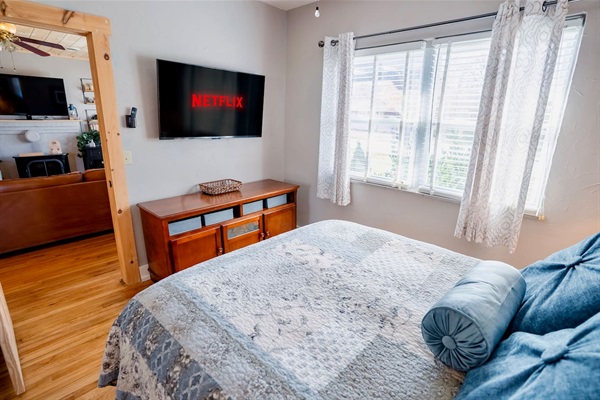 Queen bedroom with a wall-mounted smart TV, bright windows, and hardwood floors. Comfortable setup with plenty of natural light and easy access to the living room.
