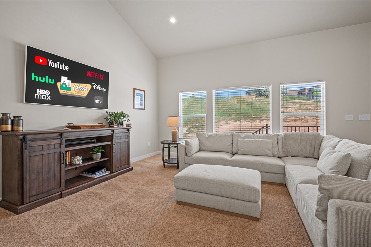 Relax in the inviting living room featuring a plush sectional, ottoman, big-screen TV for streaming, and a trio of windows that fill the space with light.