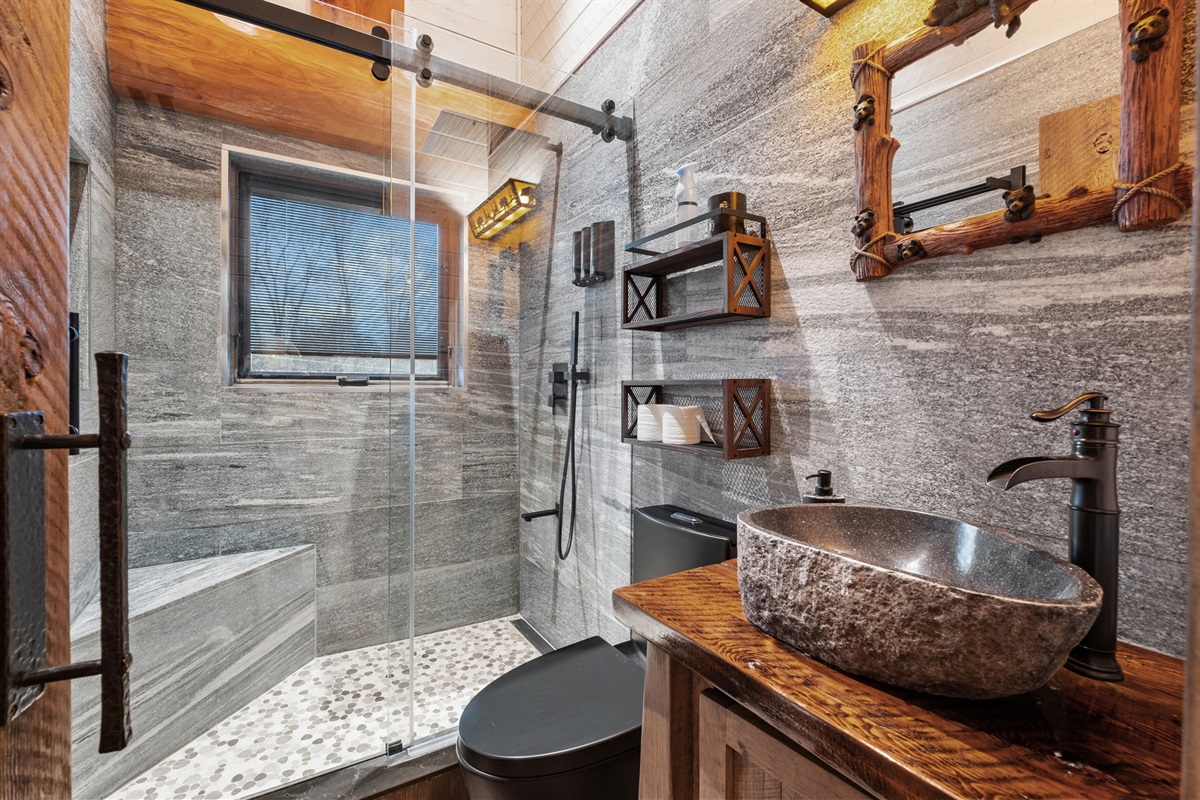 Luxury spa-style bathroom featuring a natural stone vessel sink, glass walk-in shower with rainfall head, and warm rustic wood finishes.