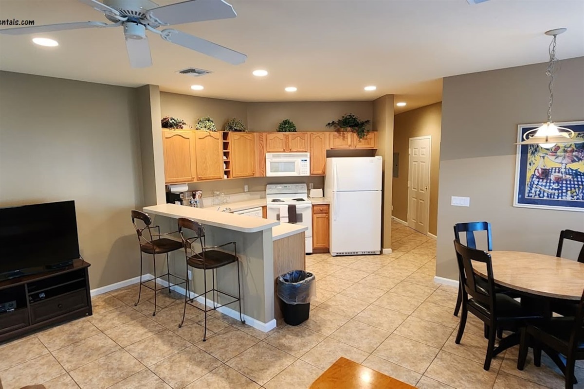 Open-concept kitchen, dining, and living area with breakfast bar seating, large TV, and tile flooring throughout.