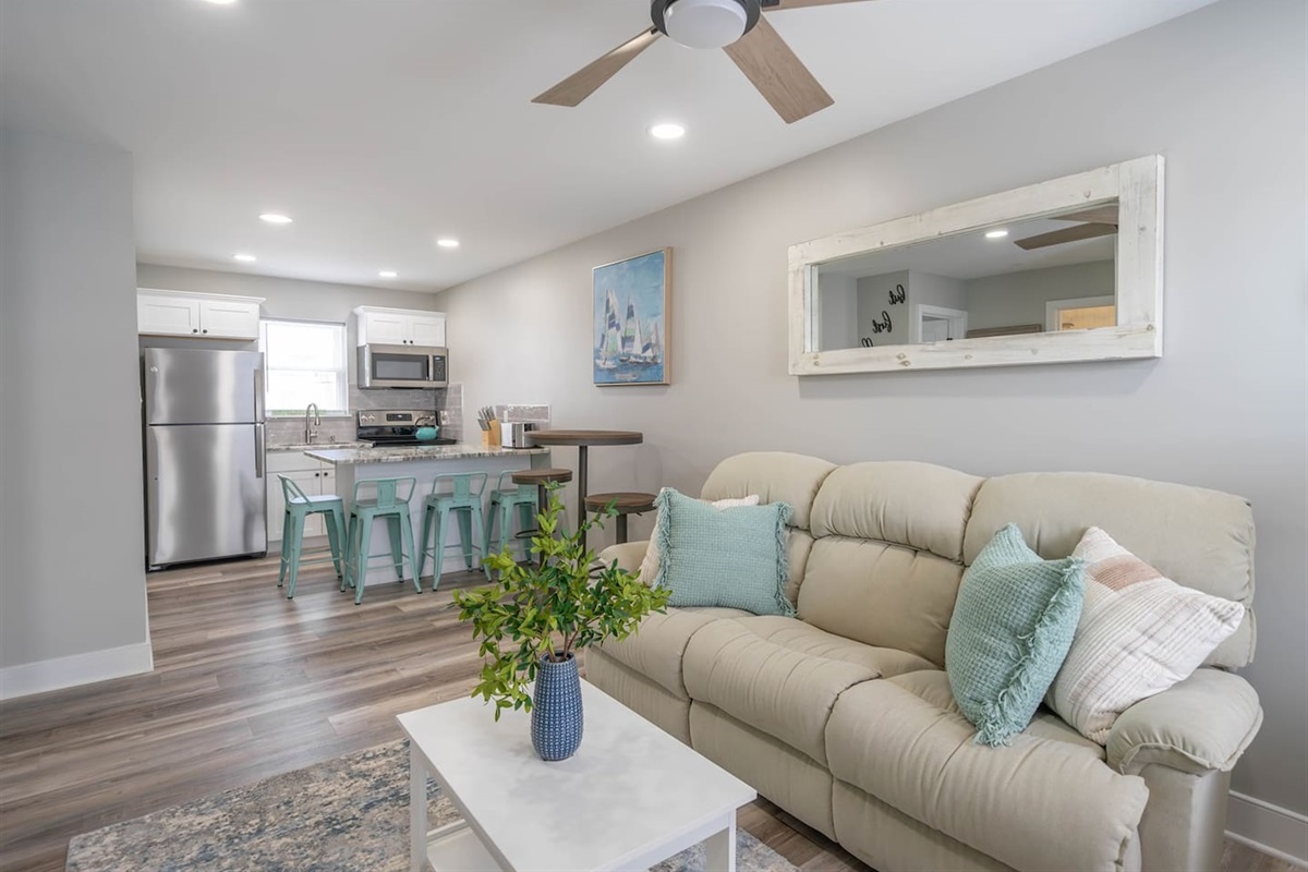 Open, airy living area flowing into the kitchen, featuring plush seating, ceiling fan, and soft coastal styling. The layout encourages relaxed lounging, easy conversation, and casual meals, making it a comfortable hub for unwinding throughout the stay.