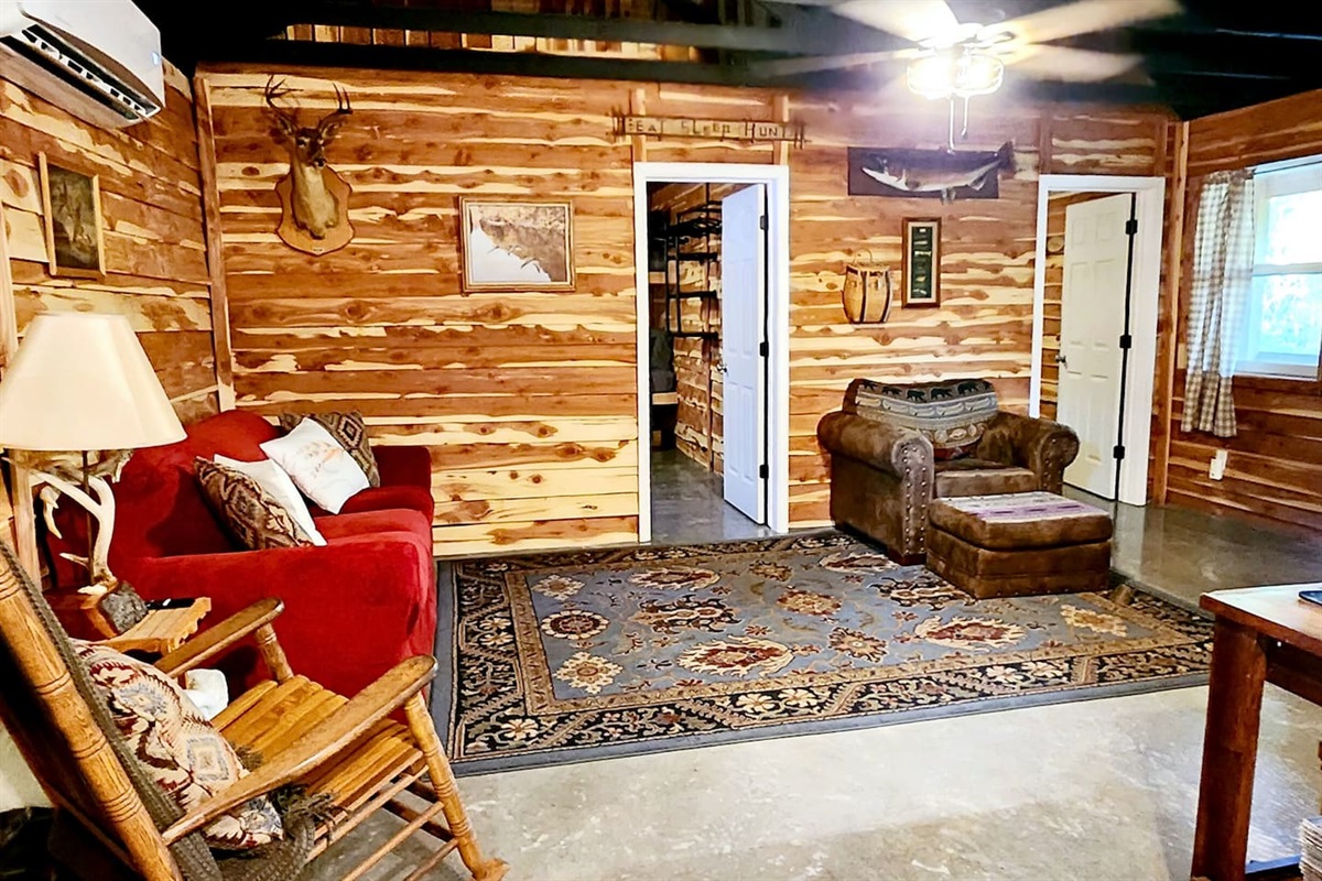 This open concept lodge living room blends rustic charm with comfort featuring a red sofa, cozy chair, rocking recliner, and forest-inspired decor. Layered rugs, wood walls, and soft lighting create a space perfect for relaxing or reconnecting