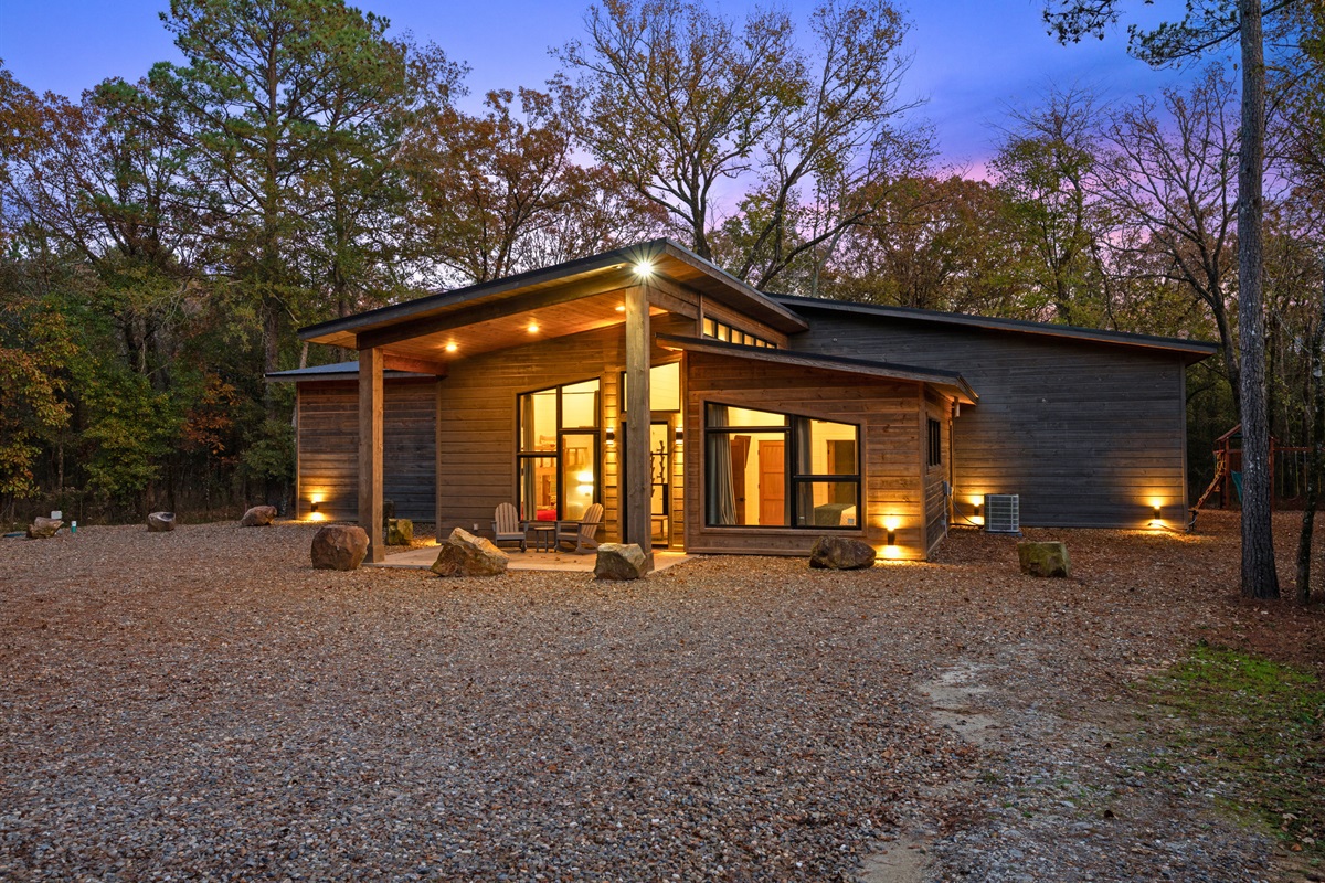 A front-facing look at the home as the sun sets—warm lights, welcoming entry, and forest charm all around.