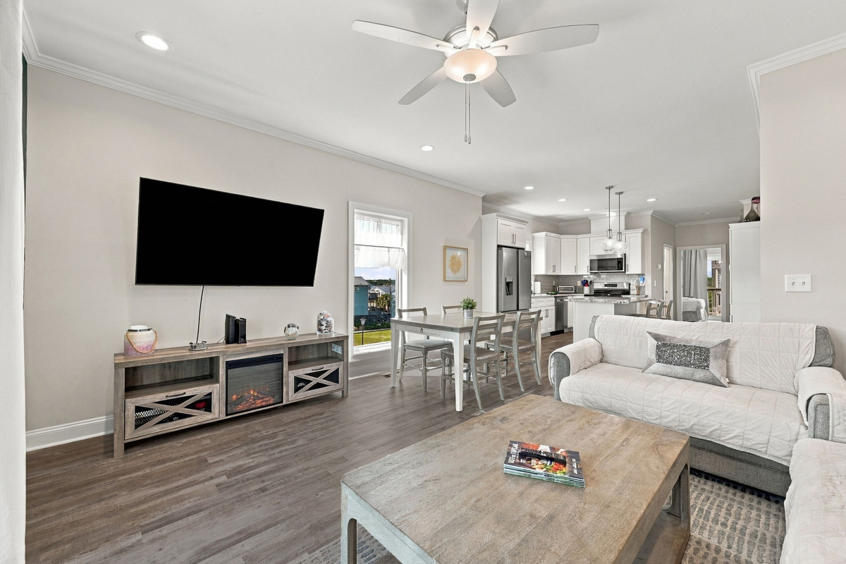 Spacious main floor layout featuring a large wall-mounted flat-screen TV over a rustic media console with an electric fireplace, flowing smoothly into the central dining area and kitchen beyond.   