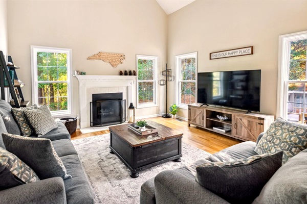 Gather in the light-filled living room with vaulted ceilings, cozy seating, and a central fireplace overlooking peaceful wooded views.