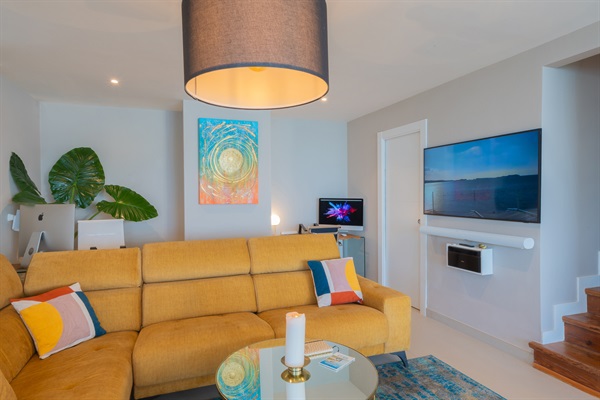 Relax in Casa Folimanka's living room, adorned with a plush sofa, elegant floor lamp, and an ultra-thin TV offering streaming services. Enhance your experience with iMacs at your disposal and a Sonos sound system featuring Atmos for immersive audio. Your e