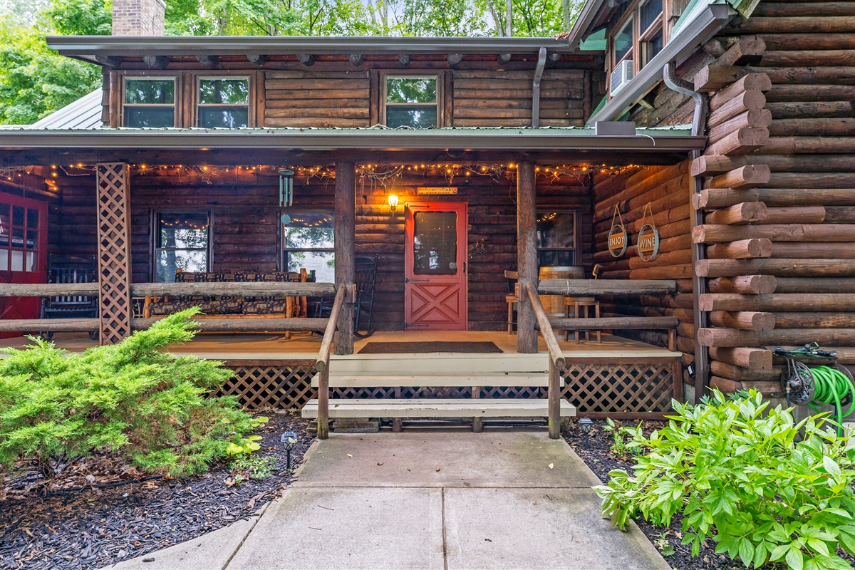 Charming log cabin entrance with a welcoming front porch, twinkling string lights, and rustic seating for cozy evenings.