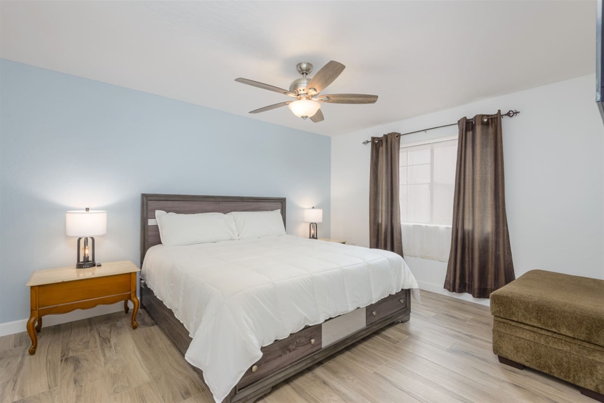 Bedroom with a clean bed setup, ceiling fan, nightstand lighting, and space to settle in.
