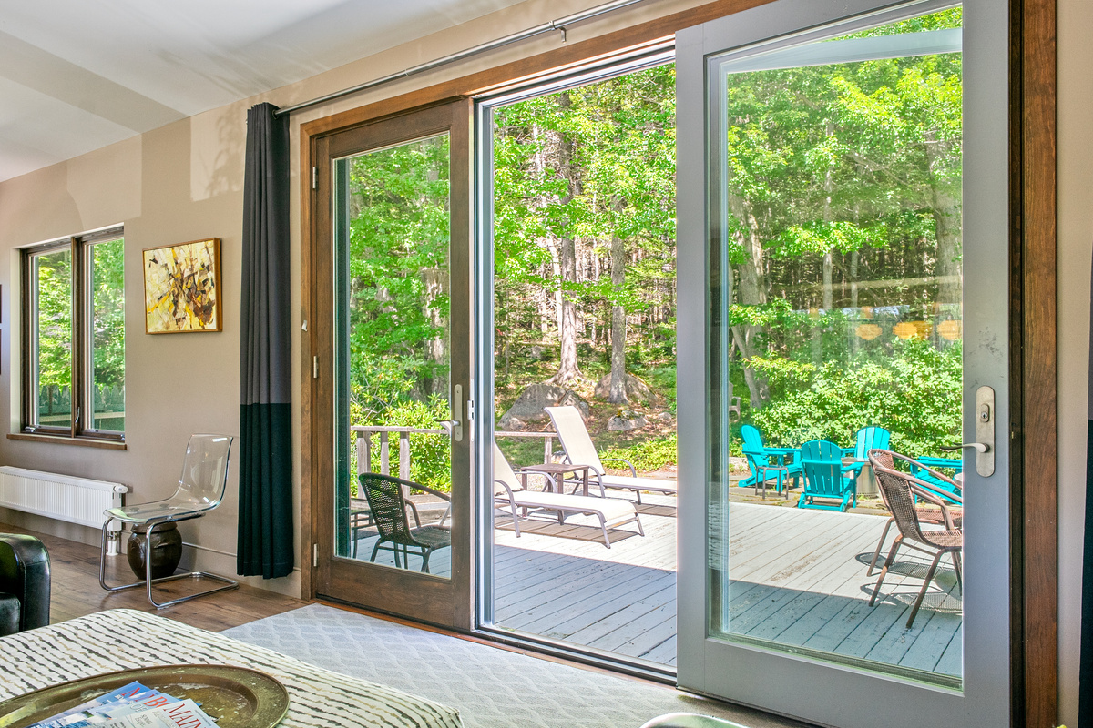 Seamless indoor-outdoor living. Premium Andersen glass doors with screens open wide to "The Crag's" back deck—outfitted with sun loungers, outdoor seating, and bright Adirondack chairs framed by crisp Maine air and Acadia woods.