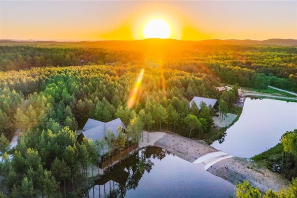 A stunning aerial view of a secluded cabin community surrounded by lush greenery, with the sun setting over the mountains. This peaceful getaway is ideal for outdoor lovers and those seeking tranquility.