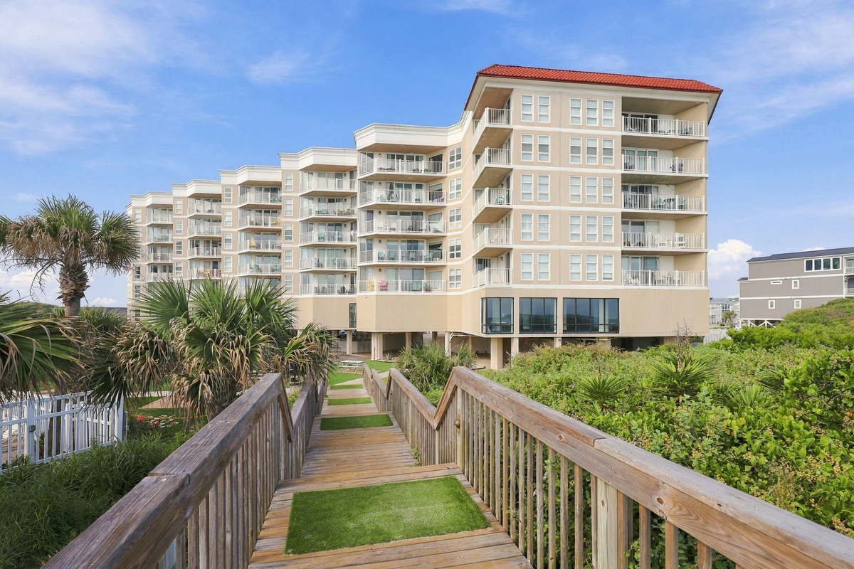 Wooden walkway leading toward the resort building, surrounded by lush green lawns and palm trees.