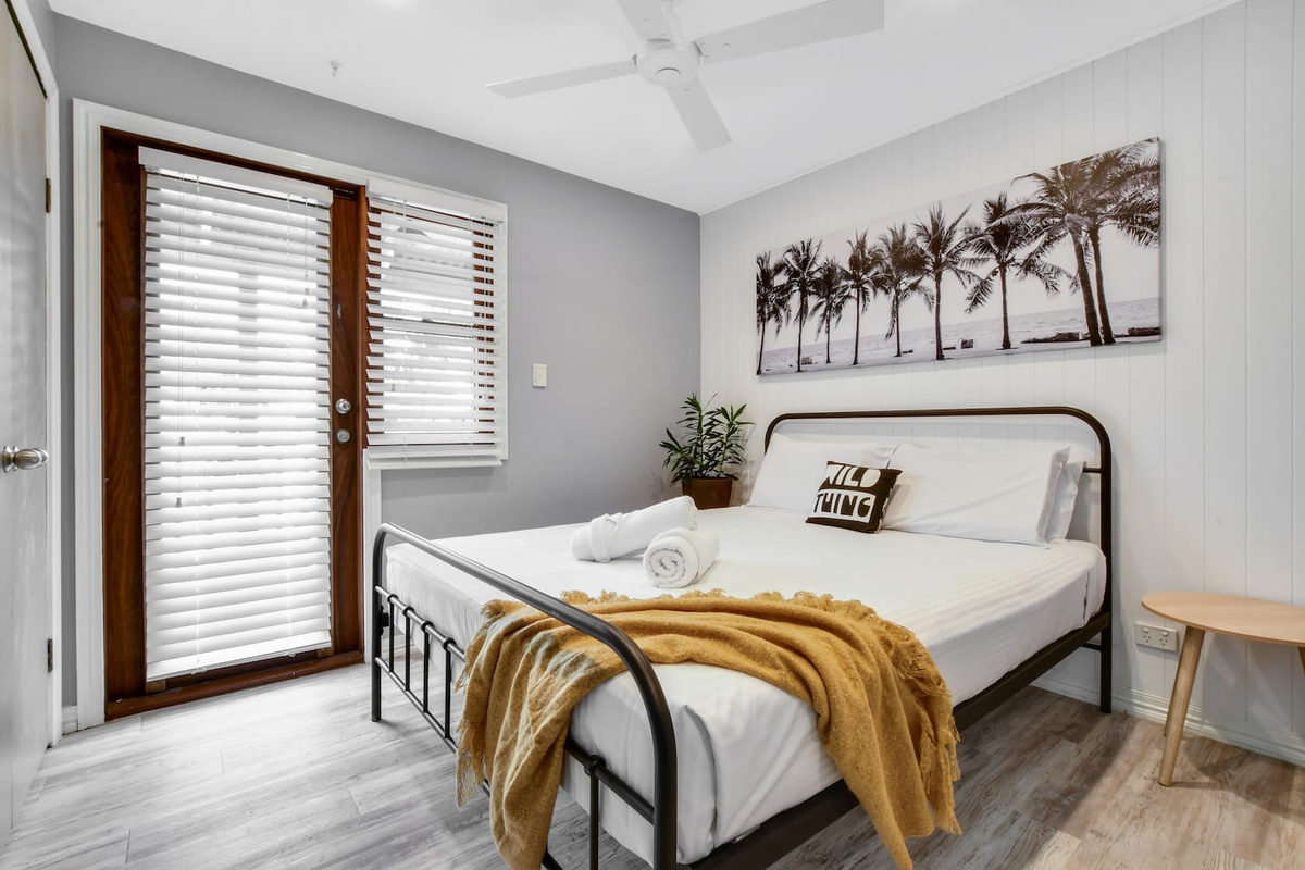 Palm artwork, timber blinds and warm coastal tones give this bedroom at Aaron's Beachside Boat House a distinctive beach-house feel.