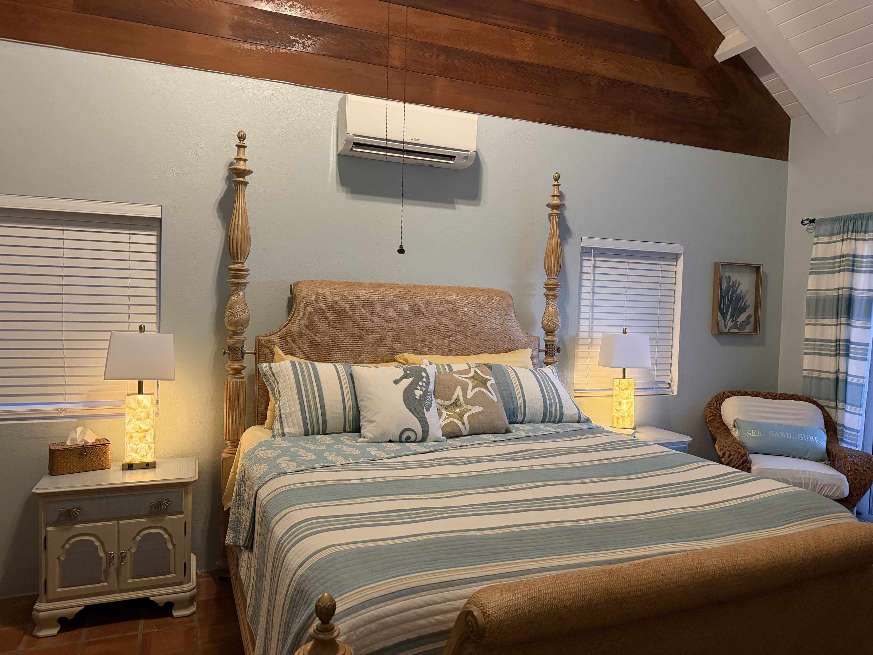 A comfortable wicker armchair sits in the corner alongside framed nautical artwork, while large sliding glass doors open directly onto the sunlit pool deck with views of the Caribbean ocean