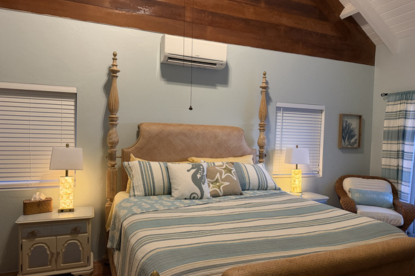 A comfortable wicker armchair sits in the corner alongside framed nautical artwork, while large sliding glass doors open directly onto the sunlit pool deck with views of the Caribbean ocean