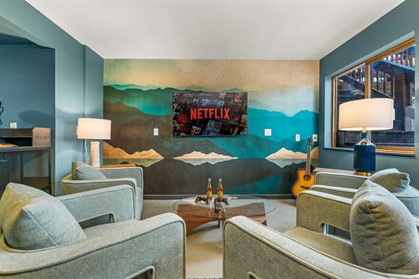 🎬🛋️ Cozy movie lounge with plush chairs, big-screen TV 📺, and a bold mountain mural—kick back with a drink 🍻 or strum a tune on the guitar 🎸✨