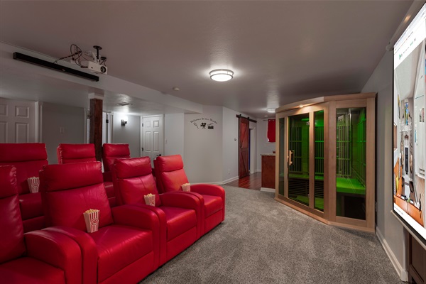 Theater-style lounge with red recliner seating, a large projector screen, popcorn setup, and an in-room sauna, perfect for movie nights and relaxing with your group.