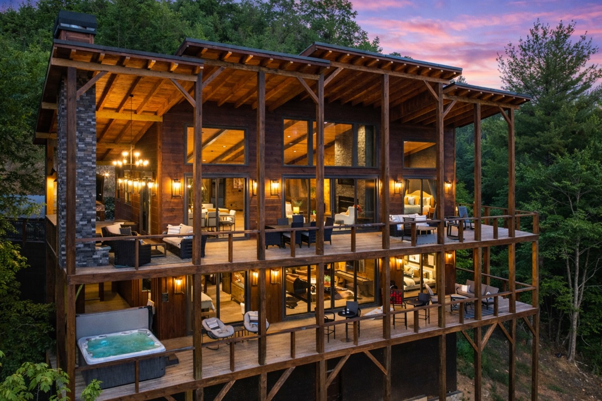 Expansive glass walls, warm ambient lighting, multiple decks with luxurious seating, a hot tub, and breathtaking forest views.