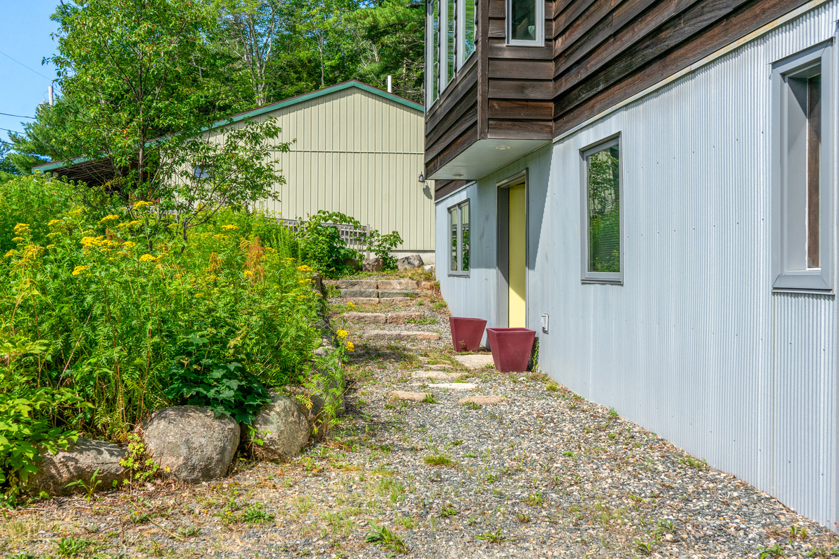 A stone-and-gravel path leads to the main Base Camp entrance of "The Crag." Framed by native greenery and granite boulders, this covered doorway welcomes guests into the ground floor of our Acadia sanctuary.