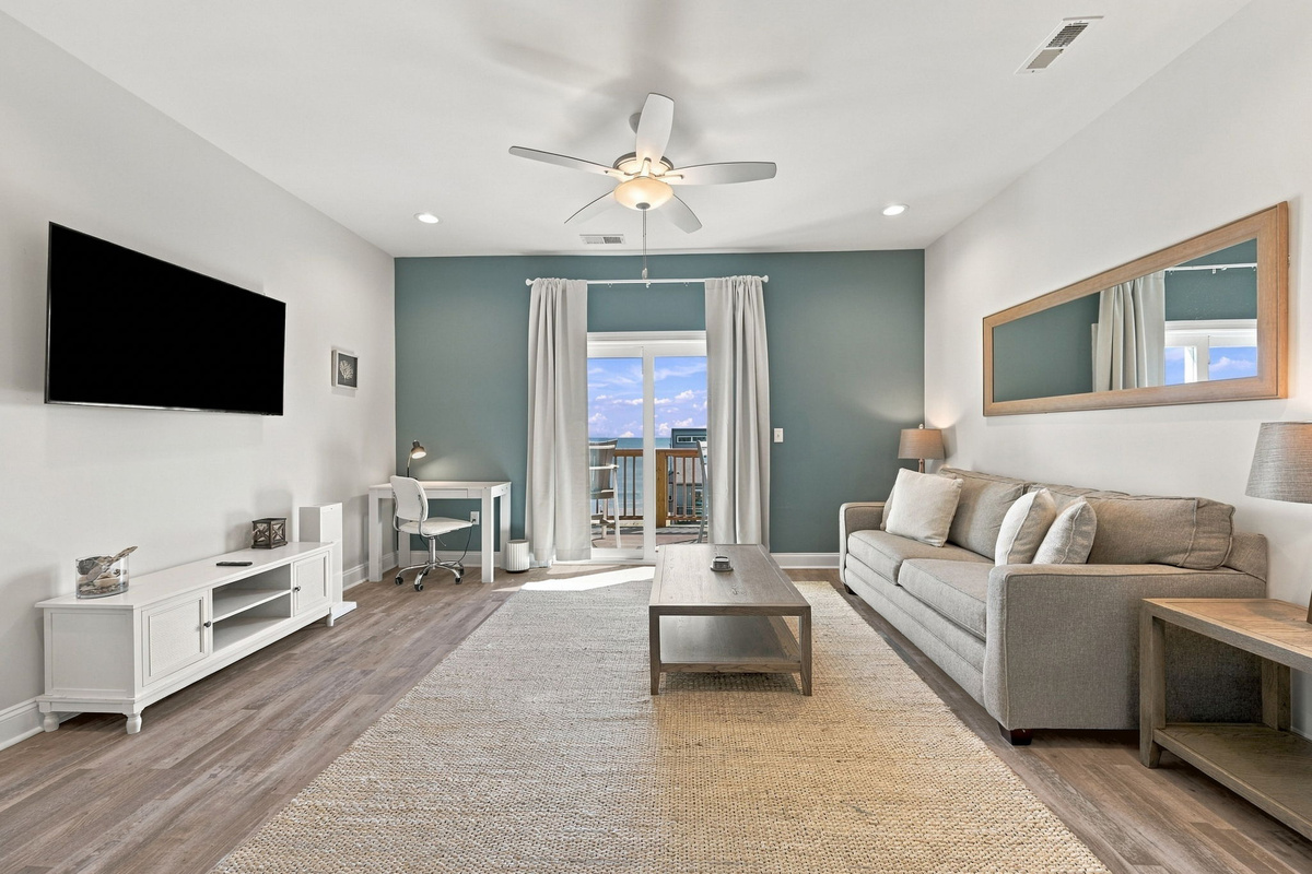 Adjoining sitting area featuring a neutral sofa, large wall-mounted flat-screen TV, dedicated work desk, and sliding glass doors opening to a private balcony with water views.   